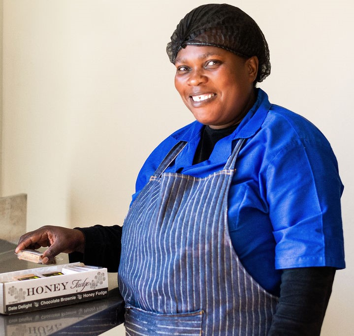 A smiling woman in a blue shirt and striped apron stands behind a counter displaying boxes of Honey Fudge, a local sweet treat.